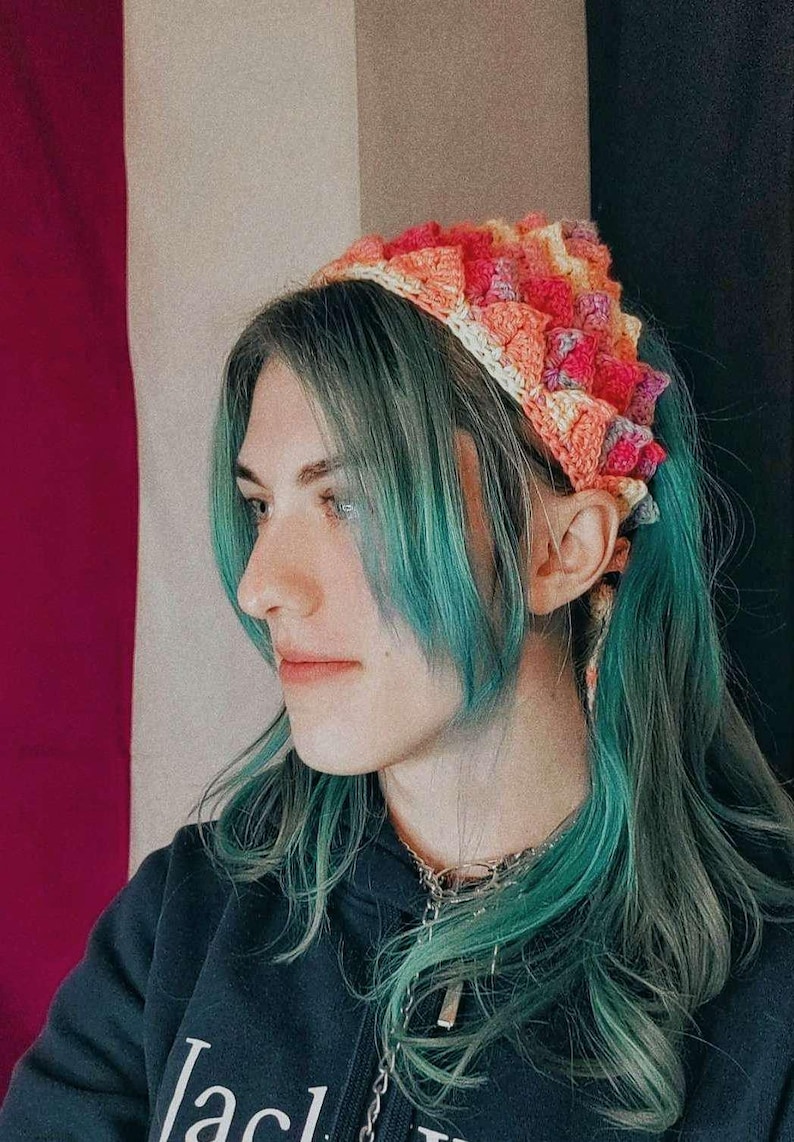K&ouml;nnte beinhalten: Eine Person mit blaugr&uuml;nem Haar tr&auml;gt ein geh&auml;keltes Stirnband in den Farben Pink, Orange und Gelb. Das Stirnband ist am Hinterkopf zu einem Knoten gebunden. Die Person tr&auml;gt einen schwarzen Kapuzenpullover mit dem Wort "Jack" auf der Vorderseite.