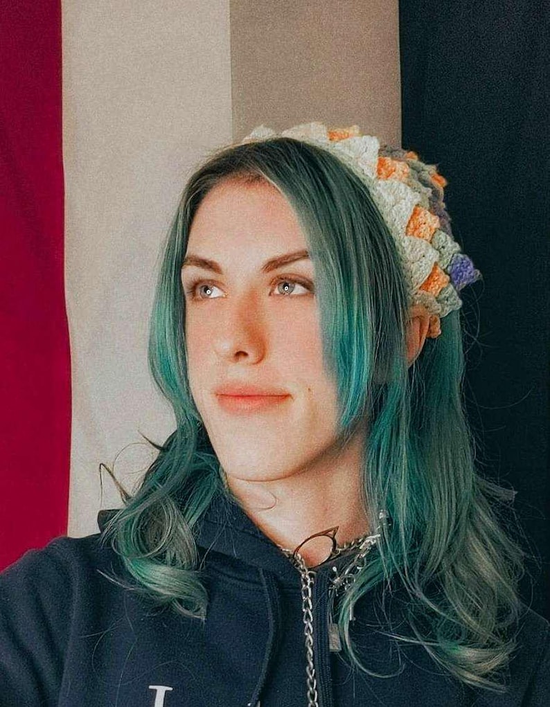 K&ouml;nnte beinhalten: Eine Person mit langen, blaugr&uuml;nen Haaren tr&auml;gt ein geh&auml;keltes Stirnband mit einem mehrfarbigen Muster. Das Stirnband besteht aus verschiedenen Gr&uuml;nt&ouml;nen, Gelb, Orange und Lila. Die Person tr&auml;gt einen schwarzen Hoodie und eine silberne Kettenkette.