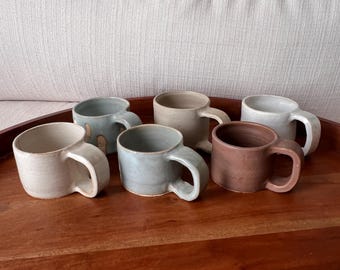 Peut inclure: Six tasses en céramique dans différentes nuances de brun, gris et blanc. Les tasses ont des poignées arrondies et sont disposées sur une surface en bois.