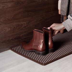 May include: Brown leather Chelsea boots with elastic side panels, placed on a patterned doormat. The boots have a polished finish and a low heel. The background includes a wooden wall and a light-colored floor.