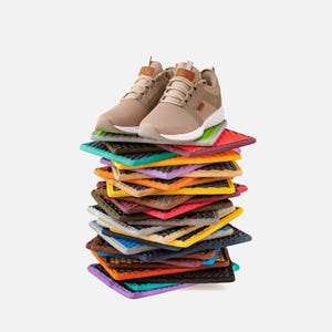 Könnte beinhalten: Ein Paar beigefarbene Sportschuhe mit weißen Sohlen und braunen Lederakzenten steht auf einem bunten Stapel quadratischer, strukturierter Matten. Die Matten sind in verschiedenen Farben gehalten, darunter Grün, Rot, Gelb und Lila.