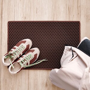 Op de afbeelding: Een bruine deurmat met een ruitpatroon, bruine en beige sneakers met groene veters en een lichtgekleurde jas en zwarte hoed op een houten vloer. De deurmat is rechthoekig en heeft een textuur.