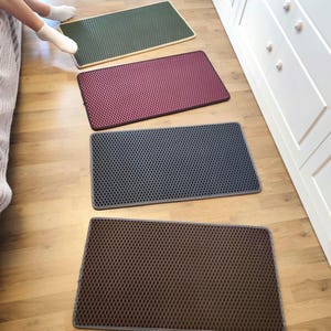 Op de afbeelding: Vier kattenmatten in verschillende kleuren, groen, rood, grijs en bruin, liggen op een houten vloer. De matten zijn gemaakt van een getextureerd materiaal met kleine gaatjes.