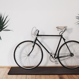 Könnte beinhalten: Ein schwarzes Fahrrad mit silbernem Lenker und einem beigefarbenen Ledersattel steht vor einer weißen Wand. Das Fahrrad steht auf einer dunkelbraunen Matte, darunter ein Holzboden. Grüne Pflanzen sind auf beiden Seiten zu sehen.