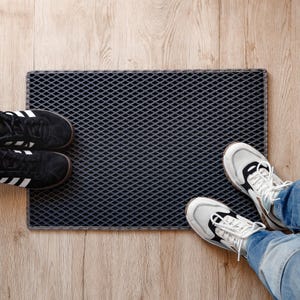 Op de afbeelding: Een zwart-witte schoen met patroon en een witte schoen met zwarte veters liggen op een donkergrijze deurmat met een ruitpatroon. De deurmat ligt op een lichtbruine houten vloer.