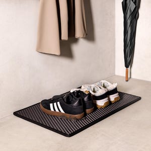 Op de afbeelding: Een zwarte rechthoekige deurmat met een geometrisch patroon, met twee paar sneakers. Eén paar is zwart met witte strepen, de andere is wit en beige. Een zwarte paraplu en jas hangen op de achtergrond.