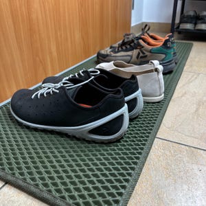 May include: A collection of shoes, including black perforated sneakers with white laces and soles, beige flats, and brown and orange athletic shoes, are arranged on a green mat.