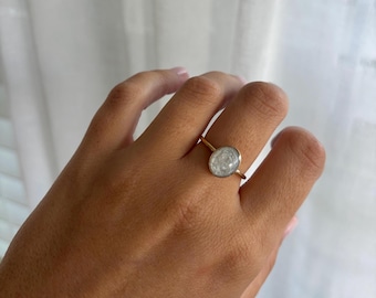 May include: A gold ring featuring an oval, light-colored stone. The ring is set on a slender gold band. The stone has a cloudy, textured appearance. The ring is shown on a person's finger.