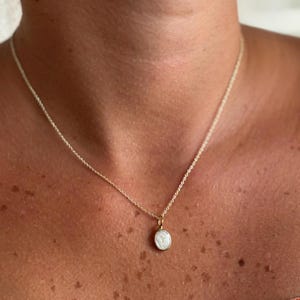 Pode incluir: Um delicado colar de ouro com um pequeno pingente redondo. O pingente apresenta um design branco dentro de uma moldura dourada. O colar é mostrado em um fundo de tom de pele, destacando os detalhes finos e a elegância da joia.