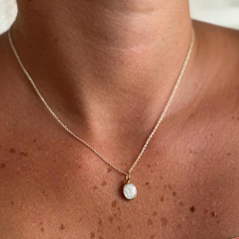 May include: A delicate gold necklace with a small, round pendant. The pendant features a white design within a gold frame. The necklace is shown against a skin tone background, highlighting the jewelry's fine details and elegant simplicity.