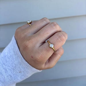 May include: Two gold rings are displayed on a hand. One ring has a clear, round stone, while the other features a small, round, white stone set in a textured band. The rings are set against a neutral background.