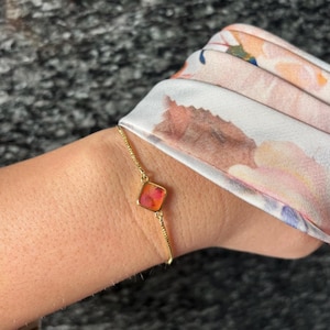Könnte beinhalten: Ein zartes Goldkettenarmband mit einem einzelnen quadratischen Anhänger in einem Farbverlauf aus Orange- und Rosatönen. Das Armband wird am Handgelenk neben einem Stoff mit weichem Blumenmuster in Pfirsich-, Weiß- und Blautönen getragen.