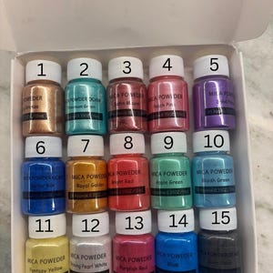 May include: A collection of 15 small jars of mica powder in various colors, including bronze, teal, mauve, peach, purple, blue, gold, red, green, and black. Each jar is labeled with a color name and volume of 0.35 oz / 10ml.