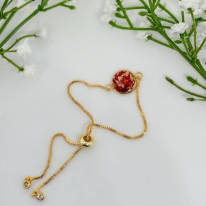 Può includere: Un braccialetto regolabile in oro con un ciondolo rotondo rosso e oro. Il ciondolo ha un aspetto simile alla resina con scaglie dorate.