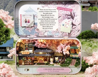 May include: A miniature dollhouse scene inside a tin box with a pink and white cherry blossom theme. The dollhouse features a small shop with a sign that reads "Flower Shop" in Japanese. There is a pink and white cherry blossom tree, a small train, and two miniature figures. The tin box lid has a drawing of a cherry blossom tree and the text "Spring Is Here" and "Cherry Blossoms in The Dreamland".