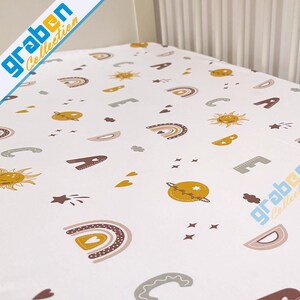 May include: White fitted crib sheet with a colorful pattern of rainbows, suns, stars, and hearts. The pattern is in shades of yellow, brown, pink, and gray.