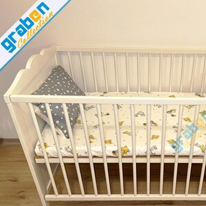 May include: A white wooden crib with a slatted design, featuring a mattress with a cartoon pattern and a gray star-patterned pillow. The crib has a scalloped headboard and footboard.