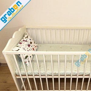 May include: White wooden crib with a white and green patterned mattress and a white pillow with a colorful animal print.