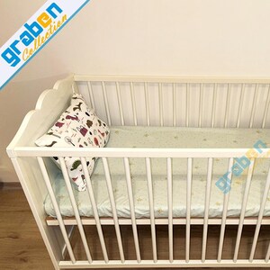 May include: White wooden crib with a white and green patterned mattress and a white pillow with a colorful animal print.