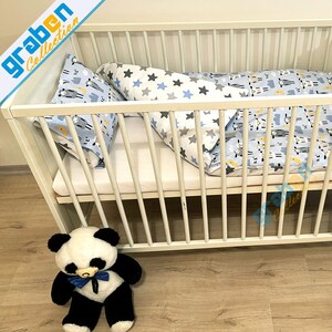 May include: White wooden crib with a blue and white patterned crib bedding set. The bedding includes a fitted sheet, a comforter, and two pillowcases. A black and white panda bear plush toy is sitting in front of the crib.