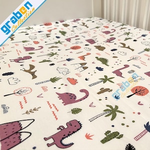 May include: A white fitted crib sheet with a dinosaur and jungle themed print. The print features pink, green, orange, and brown dinosaurs, palm trees, and mountains.