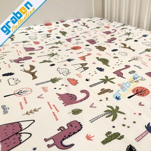 May include: White fitted crib sheet with a dinosaur and jungle themed print. The print features pink, green, orange, and brown dinosaurs, palm trees, mountains, and other jungle elements.