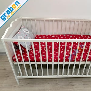 May include: White wooden crib with a red patterned fitted sheet and a gray pillow. The fitted sheet has a white pattern of snowflakes and other shapes. The pillow has a pattern of hot air balloons and other shapes. The crib has the text "graben Collection".