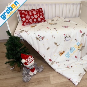 Puede incluir: Una cuna blanca con un juego de cama con temática navideña. El juego incluye una funda nórdica y una almohada roja con copos de nieve blancos. La funda nórdica presenta ilustraciones de animales con gorros de Papá Noel.