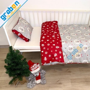 May include: A white crib with a gray and red Christmas-themed bedding set. The set includes a pillow, a blanket with a snowman and tree pattern, and a red blanket with snowflakes and Santa Claus. A small Christmas tree and a teddy bear are also present.