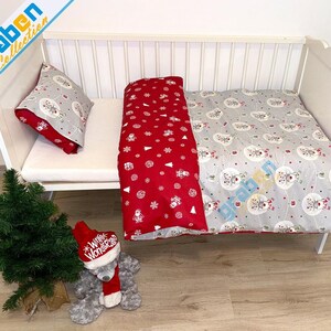 May include: A white crib with a red and gray Christmas themed bedding set. The bedding features a pattern of snowflakes, Christmas trees, and snowmen. A gray teddy bear with a red Santa hat is sitting on the floor in front of the crib.