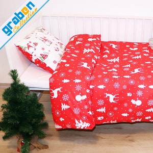 May include: Red and white Christmas themed bedding set with a white deer, snowflake, and tree pattern. The bedding is on a white crib with a stuffed animal wearing a Santa hat.
