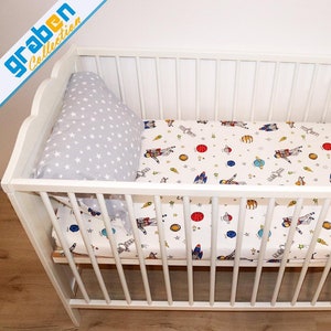 May include: White wooden crib with a white and gray star patterned fitted sheet and a gray pillow with white stars. The sheet has a colorful space-themed print with astronauts, rockets, and planets.