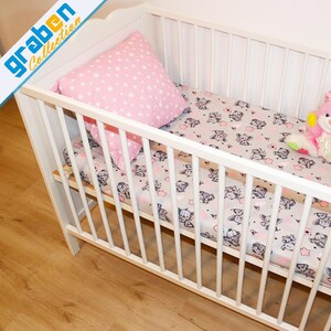 May include: White wooden crib with a pink and gray patterned sheet and a pink stuffed bunny toy. The crib has a white wooden frame and a white mattress. The sheet has a pattern of gray elephants and pink stars. The bunny toy is sitting on the sheet.