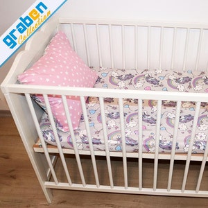 May include: White crib with a pink and white star patterned pillow and a fitted sheet with a unicorn and rainbow print.