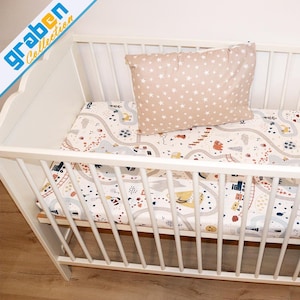 May include: A white crib with a mattress and pillow. The mattress has a road map design with cars, trucks, and buildings. The pillow is tan with white stars. The crib has a slatted design.