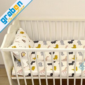May include: White wooden crib with a white and yellow dinosaur print fitted sheet and a white pillow with a rainbow and star print.