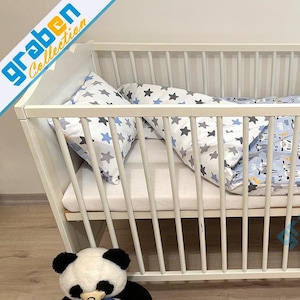 May include: White wooden crib with a white mattress and blue and gray star patterned bedding. A black and white panda bear plush toy is sitting in front of the crib.