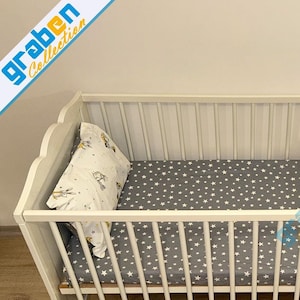 May include: A white wooden crib with a gray star-patterned sheet and a pillow with a cartoon design. The crib has a slatted design and a curved headboard. The image includes the text "graben Collection".
