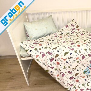May include: A white crib with a white and green dinosaur print bedding set. The bedding set includes a fitted sheet, a duvet cover, and a pillowcase. The duvet cover has a white background with a repeating pattern of green dinosaurs, palm trees, and clouds.