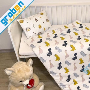 May include: A white crib with a white and yellow dinosaur patterned bedding set. The bedding set includes a pillow with a rainbow, sun, and letters on it. A beige teddy bear with a red heart is sitting on the floor next to the crib.