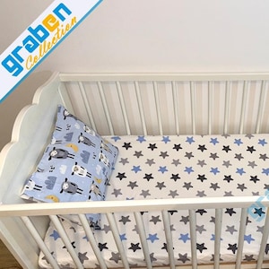 May include: A white crib with a star-patterned sheet in blue, gray, and white. A matching pillow with sheep and cloud designs rests inside. The crib has a scalloped headboard and side rails.