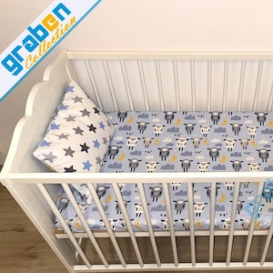 May include: White wooden crib with a blue and white sheep print fitted sheet, a gray and white star print pillow, and a white stuffed animal with antlers.