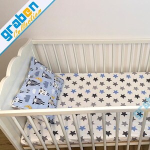 May include: White wooden crib with a white and blue star patterned fitted sheet and a blue and white pillow with a sheep and moon design. The crib has a wooden slatted base and a white painted finish.