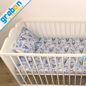 May include: White wooden crib with a blue and white sheep print fitted sheet and pillowcase. A white stuffed animal is sitting on the sheet. The crib is on a wooden floor.