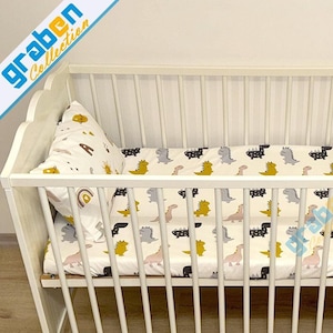 May include: White wooden crib with a white and yellow dinosaur print fitted sheet and a white pillow with a rainbow and sun print.