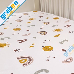 May include: White fitted crib sheet with a colorful pattern of suns, rainbows, hearts, and stars. The pattern is in shades of yellow, orange, pink, brown, and gray.