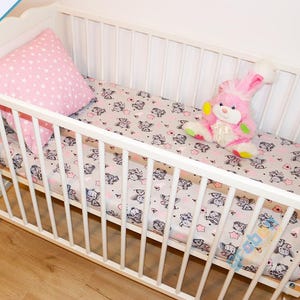 May include: White crib with a pink and white patterned sheet and a pink stuffed bunny. The crib has a white wooden frame and a white mattress.