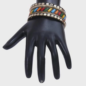 May include: A pair of gold-toned bangles with colorful rectangular inlays of red, orange, yellow, green, and blue. The bangles are adorned with small, evenly spaced circular accents. The jewelry is displayed on a black hand.