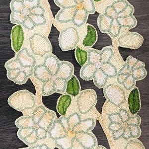 May include: A white and green beaded table runner with a floral design. The runner is made of small beads that are sewn together to create a delicate and intricate pattern. The flowers are in a variety of sizes and shapes, and they are arranged in a symmetrical pattern. The runner is perfect for adding a touch of elegance to any table.