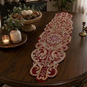 May include: A long, narrow table runner featuring a deep red fabric base with intricate gold and silver metallic embroidery. The design includes ornate scrollwork, floral motifs, and a scalloped edge, creating a detailed, symmetrical pattern.